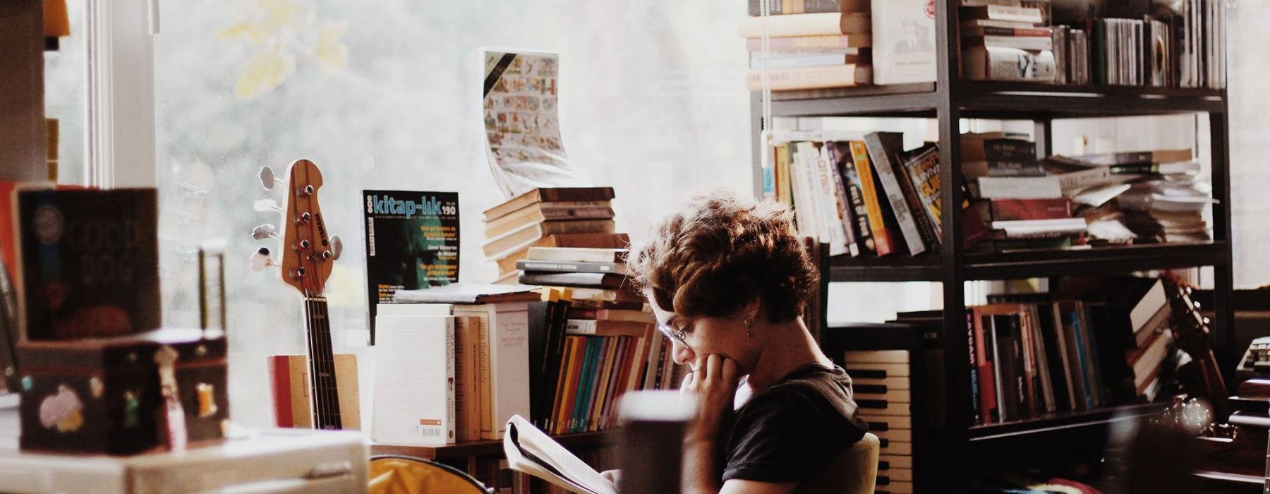 woman, surrounded by books, sits and reads in front of a picture window